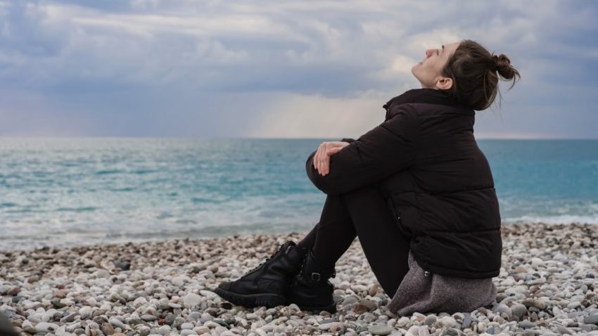 A woman sits peacefully on a rocky beach, gazing at the ocean under a cloudy winter sky.